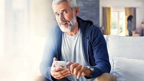 Älterer Mann in Freizeitkleidung sitzt auf Sofa und hält Smartphone in der Hand.