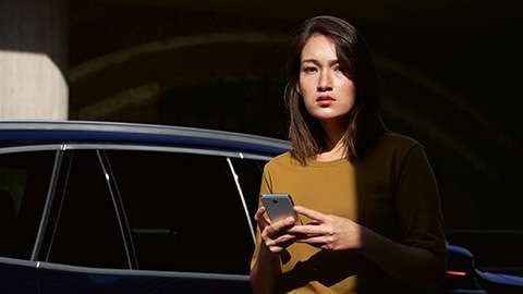 Frau mit Smartphone vor einem blauen Auto im Schatten.