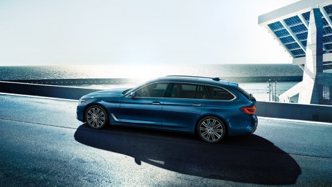 Blauer BMW Kombi auf einer Küstenstraße bei Sonnenschein, im Hintergrund das Meer und moderne Architektur.