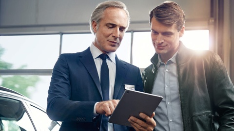 Zwei Männer betrachten ein Tablet neben einem Auto in einem hellen Raum.