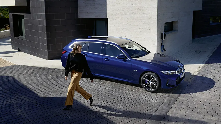 Eine Frau geht auf gepflastertem Weg neben einem blauen BMW Kombi vor einem modernen Gebäude.