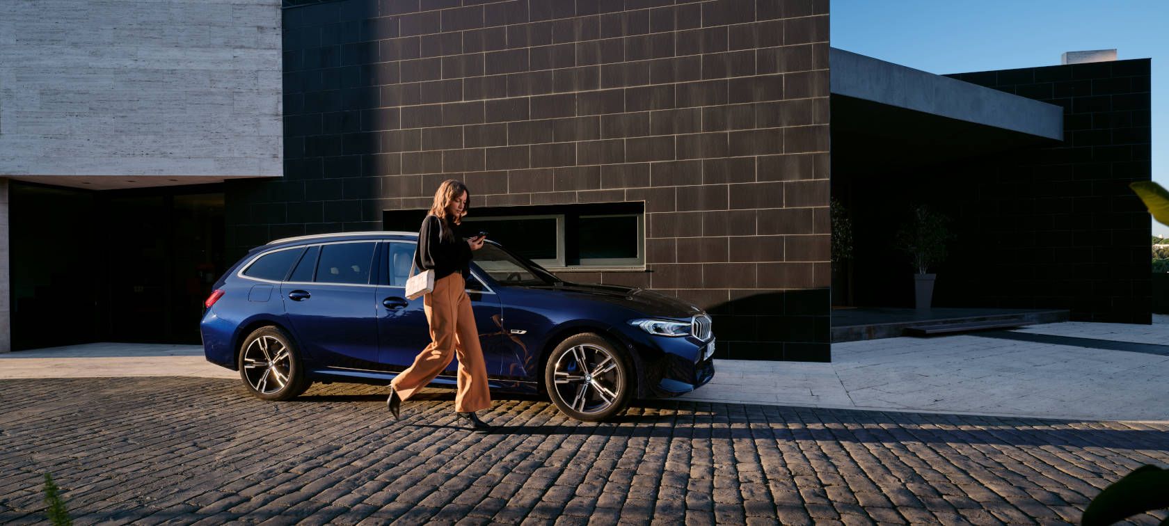 Frau geht an einem blauen BMW auf gepflasterter Auffahrt vor einem modernen Gebäude vorbei.
