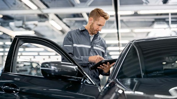 Mann in grauer Arbeitskleidung inspiziert schwarzen BMW mit Tablet in heller Werkstatt.