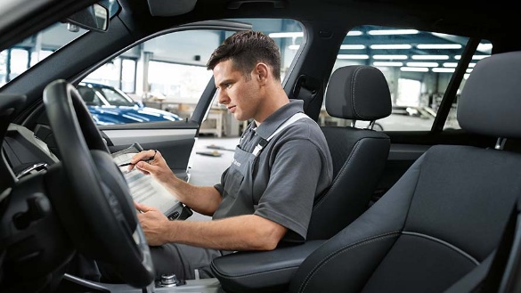 Mann in grauer Arbeitskleidung sitzt im Auto und notiert etwas, Werkstatt im Hintergrund.