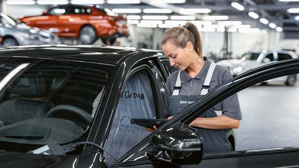 Frau in Werkstattkleidung überprüft ein schwarzes Auto mit einem Tablet in einer Werkstatt.