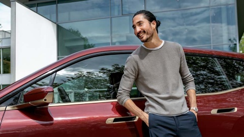 Mann lehnt an rotem BMW vor moderner Glasfassade, lächelt und schaut zur Seite.