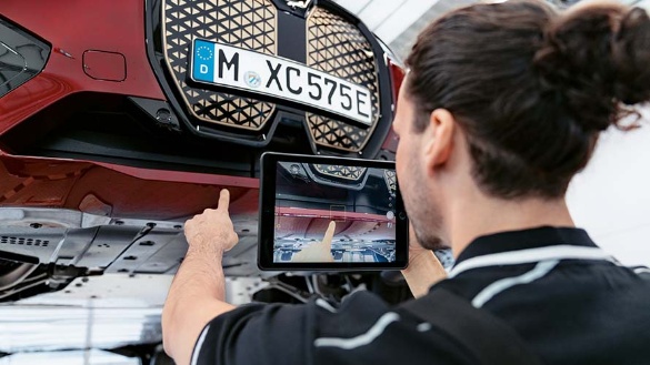 Person inspiziert mit Tablet die Unterseite eines aufgebockten roten BMWs in Werkstattumgebung.