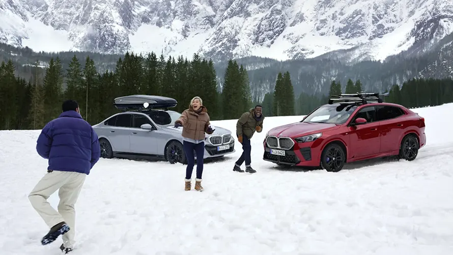 Drei Personen im Schnee vor zwei BMWs, Berge im Hintergrund.