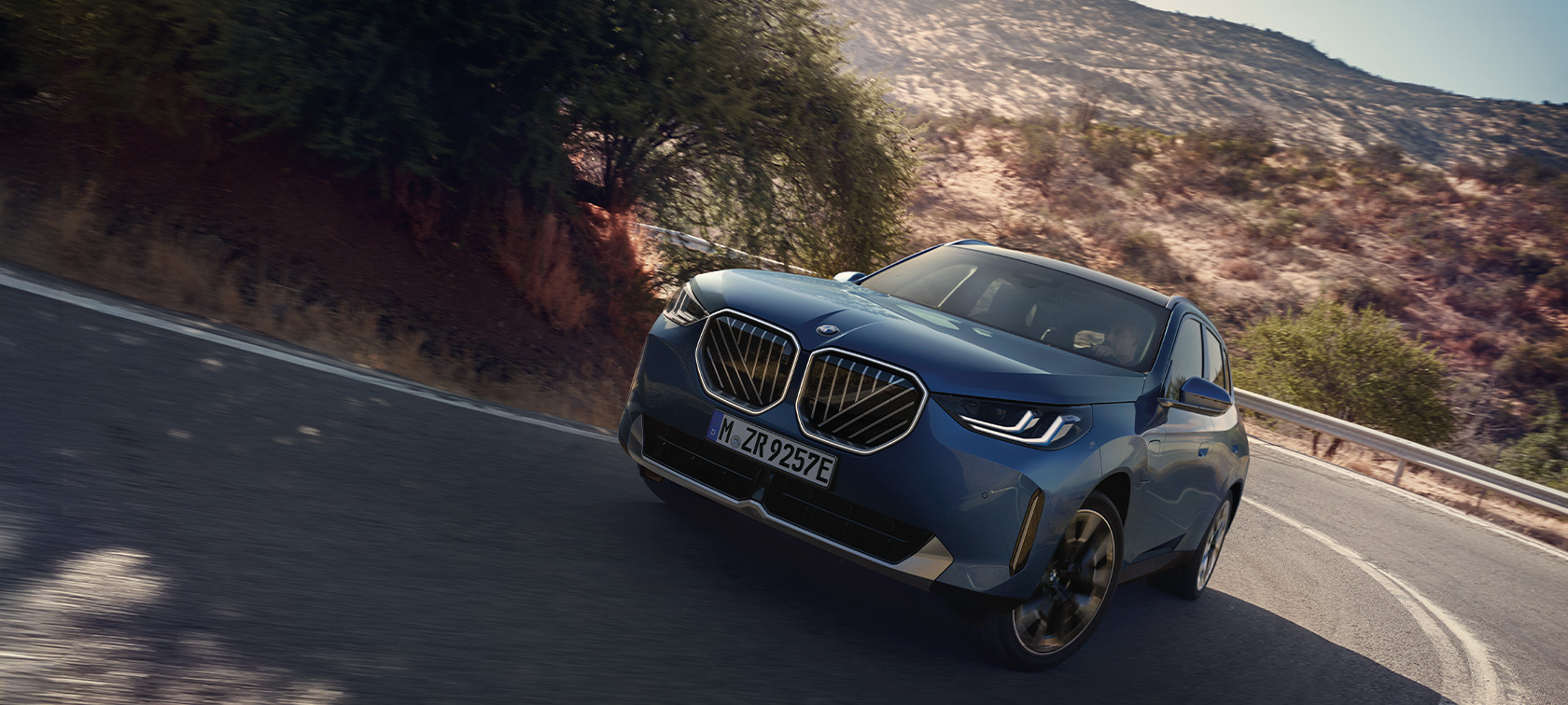 Blauer BMW fährt auf kurviger Straße, umgeben von Bäumen und Hügeln bei sonnigem Wetter.