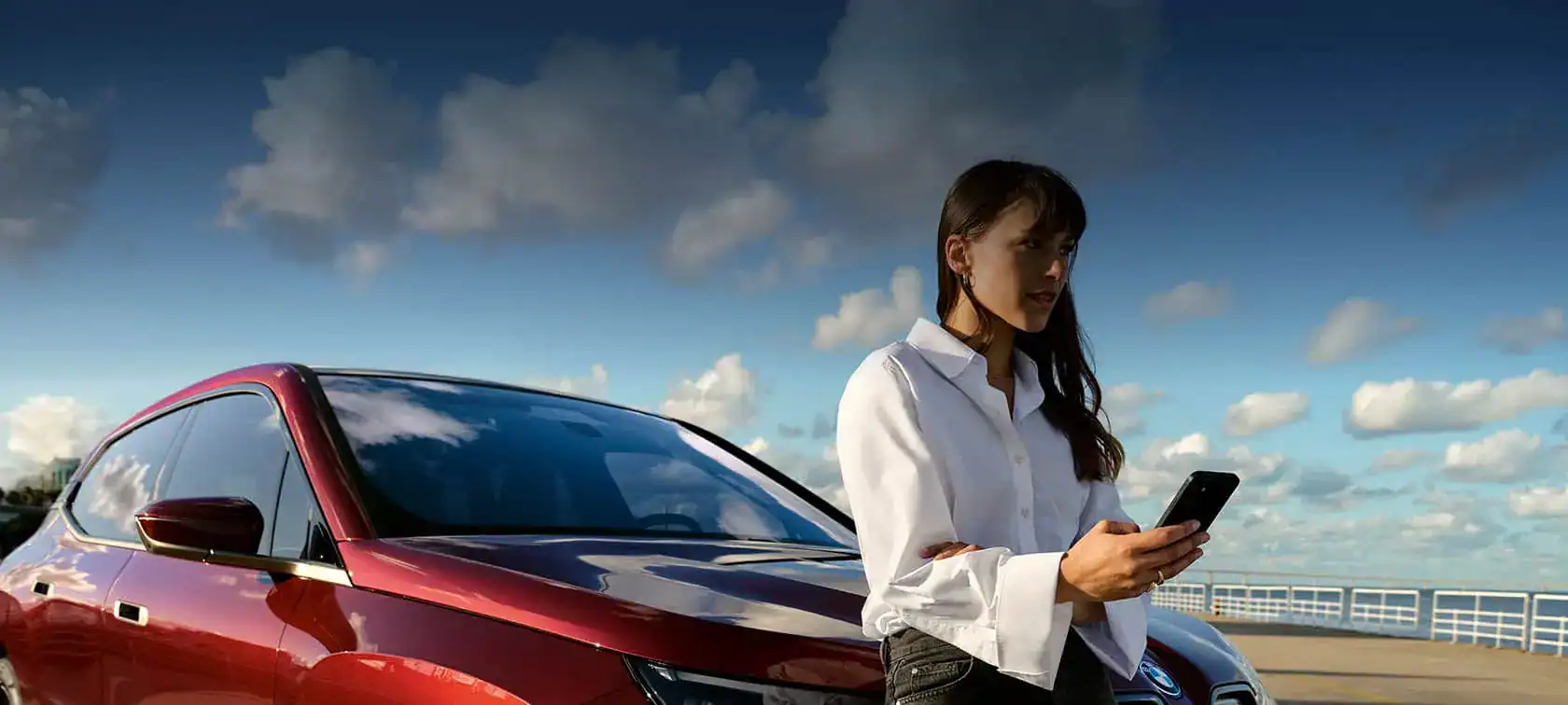 Frau mit Smartphone vor rotem BMW, blauer Himmel mit Wolken im Hintergrund.