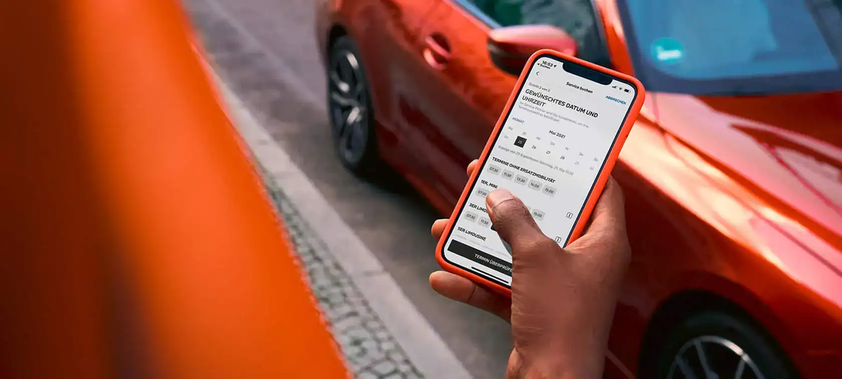Person hält Smartphone neben rotem Auto; Terminbuchung auf dem Bildschirm sichtbar.