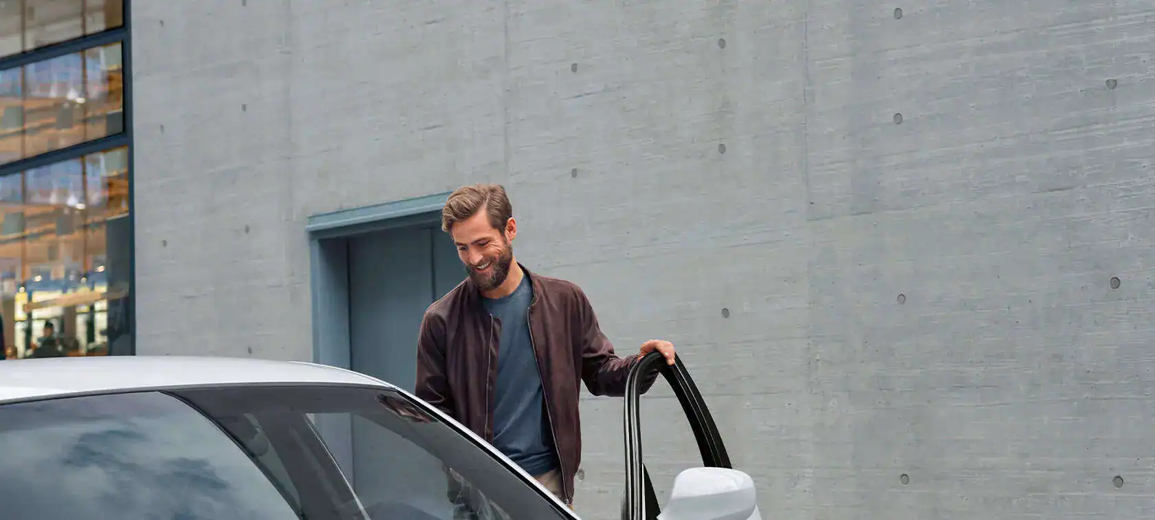 Mann öffnet Autotür vor grauer Wand, lächelt. Gebäude mit Fenstern im Hintergrund.