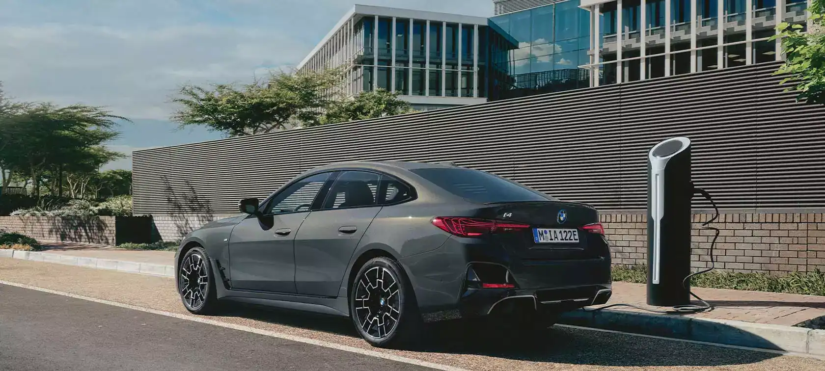 Grauer BMW i4 parkt neben einer Ladestation vor einem modernen Gebäude mit Glasfassade.