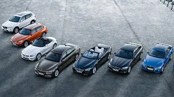 Sieben BMW-Fahrzeuge in verschiedenen Farben nebeneinander auf grauem Asphalt.