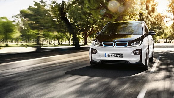 Weißer BMW i3 fährt auf einer von Bäumen gesäumten Straße bei Sonnenschein.