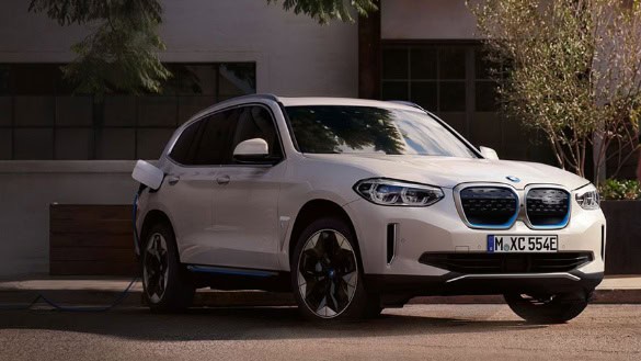 Weißer BMW iX3 steht vor einem Gebäude, an ein Ladekabel angeschlossen.